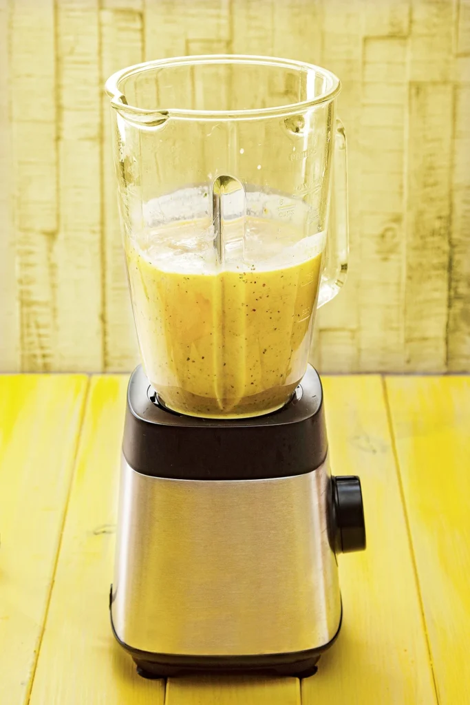 A blender stands on a yellow wooden underground. The glass container is filled with the mango popsicle mixture.