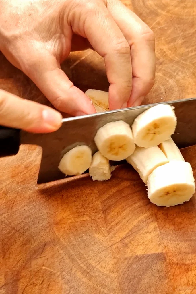 One hand is holding a banana, the other one a huge knife that chop the banana on a wooden cutting board.