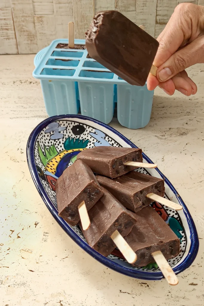 Behind an oval bowl of five chocolate popsicles, a hand holds a chocolate popsicle just removed from the popsicle mold.