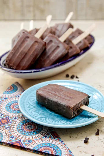 There is a chocolate popsicle on a blue plate. Next to it is a patterned napkin. Behind it is a long bowl with a pile of popsicles. Chocolate pieces lie around on the stone base.