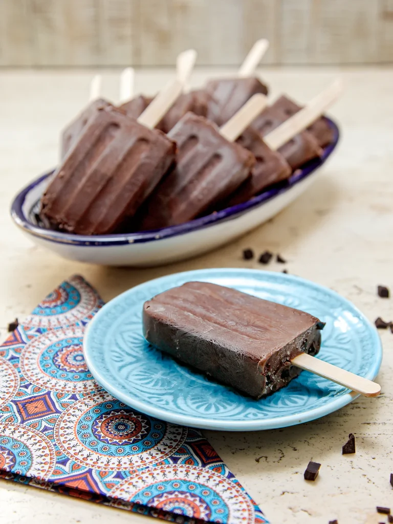 There is a chocolate popsicle on a blue plate. Next to it is a patterned napkin. Behind it is a long bowl with a pile of popsicles. Chocolate pieces lie around on the stone base.