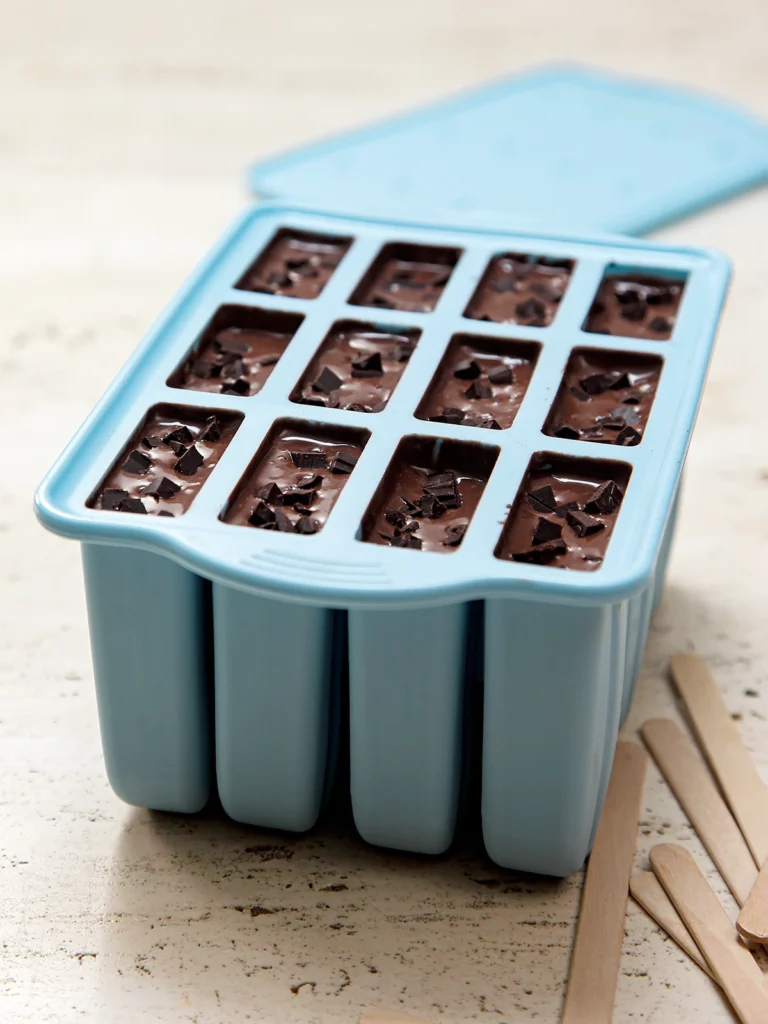 Chocolate mixture is filled into the popsicle mold with small chocolate pieces on top, wooden sticks in front, and the lid in the background.