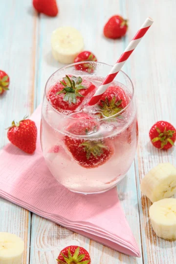 A glass of water with fruit ice cubes and a straw stands on a light blue wooden base. Next to the glass are slices of bananas, strawberries and a napkin.