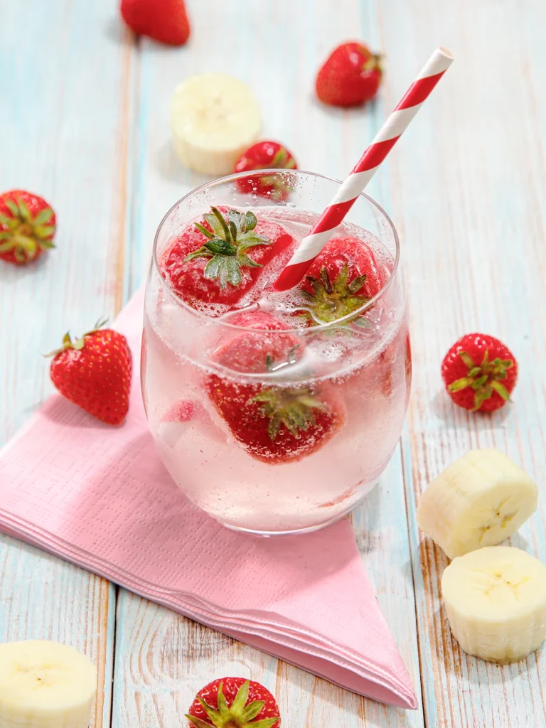 A glass of water with fruit ice cubes and a straw stands on a light blue wooden base. Next to the glass are slices of bananas, strawberries and a napkin.