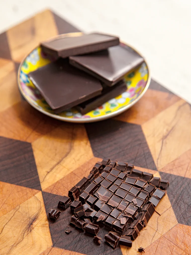 Finely chopped chocolate lies on a patterned wooden board. Also on the board is a small bowl with whole pieces of chocolate.