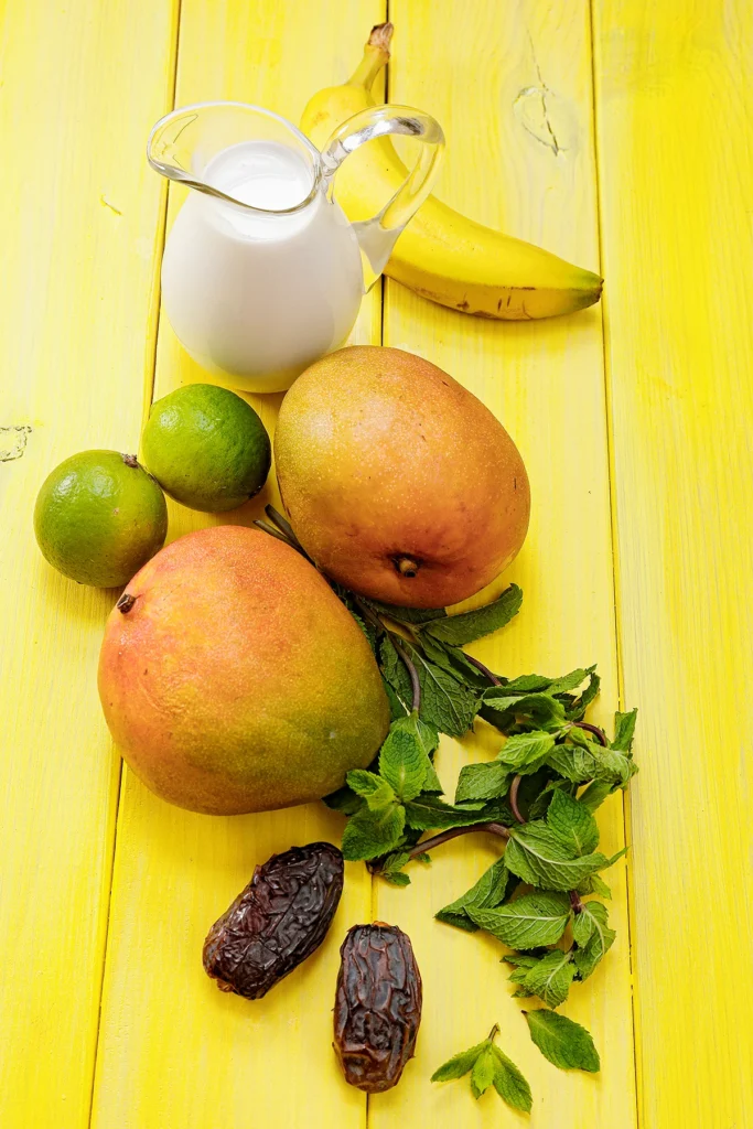 On a yellow wooden underground are lying two mangos, two lemon, two dates one banana and fresh mint leaves. Behind thte mangos stands a jug with coconut milk.