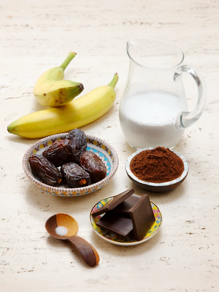 Three bowls stand on a travertine stone base. One with dates, chocolate pieces and cocoa. In the foreground, a wooden spoon with salt.