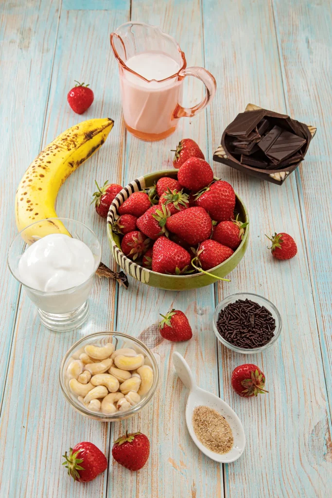 There are many bowls on a light blue wooden background. In a large one in the middle are fresh strawberries, surrounded by others with chocolate, chocolate sprinkles, vanilla sugar, soaked cashew nuts, a jar of coconut yogurt, a ripe bannana and a jug of coconut milk. A few strawberries are scattered in between.