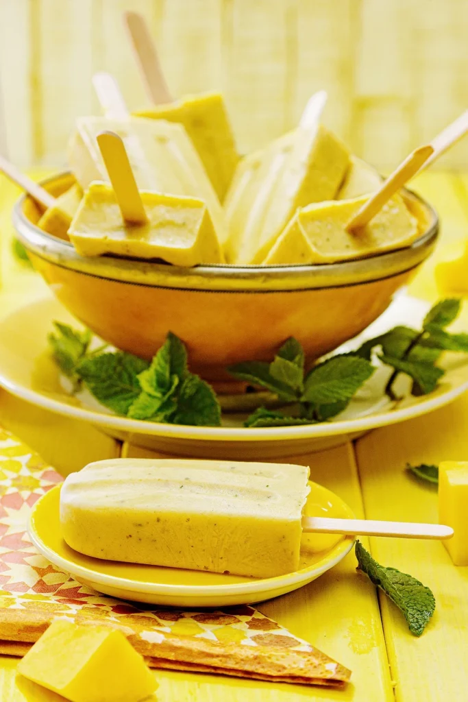 A small yellow plate with a mango mint popsicle lies on a yellow wooden background in the foreground. The plate is placed on a patterned napkin. Behind it is a plate with a large yellow bowl on it, filled with more mango mint popsicles. On the edge of the plate are tufts of mint leaves. In the foreground, pieces of mango lie on the wooden background.