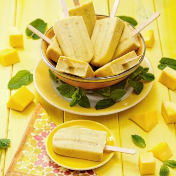 A small yellow plate with a mango mint popsicle lies on a yellow wooden background in the foreground. The plate is placed on a patterned napkin. Behind it is a plate with a large yellow bowl on it, filled with more mango mint popsicles. On the edge of the plate are tufts of mint leaves. Pieces of mangos and mint leaves are scattered on the wooden underground.