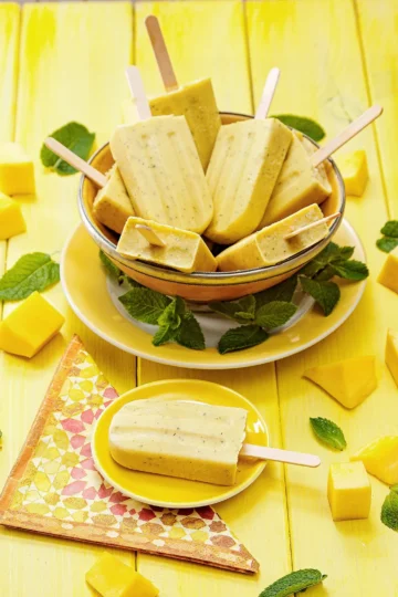 A small yellow plate with a mango mint popsicle lies on a yellow wooden background in the foreground. The plate is placed on a patterned napkin. Behind it is a plate with a large yellow bowl on it, filled with more mango mint popsicles. On the edge of the plate are tufts of mint leaves. Pieces of mangos and mint leaves are scattered on the wooden underground.