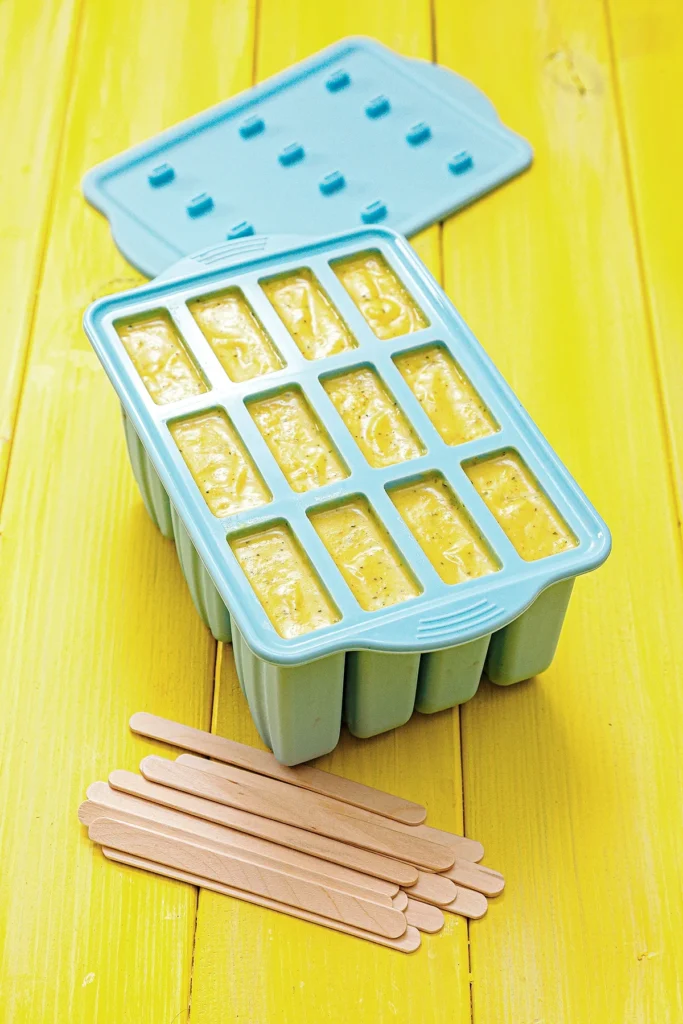 The silikon popsicle mold is filled with the mango mint mixture. The lid lies behind. In the foreground are wooden posicle sticks. The blue mold stands on a yellow wooden underground.