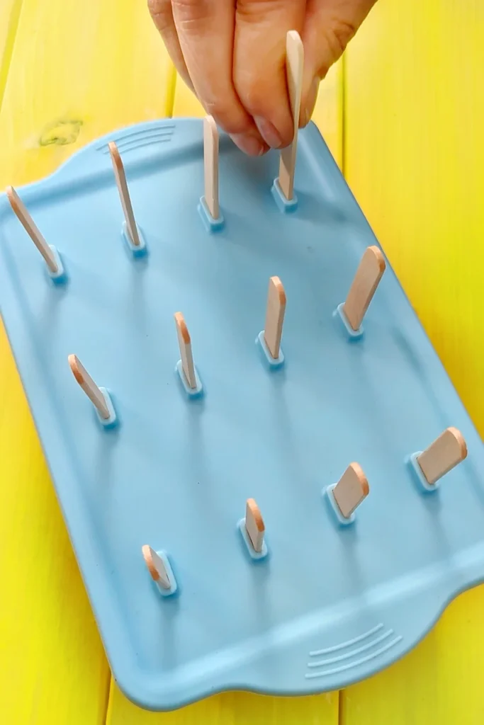 The silikon popsicle mold is closed with a lid and wooden sticks are inserted by hand. The blue mold stands on a yellow wooden underground.