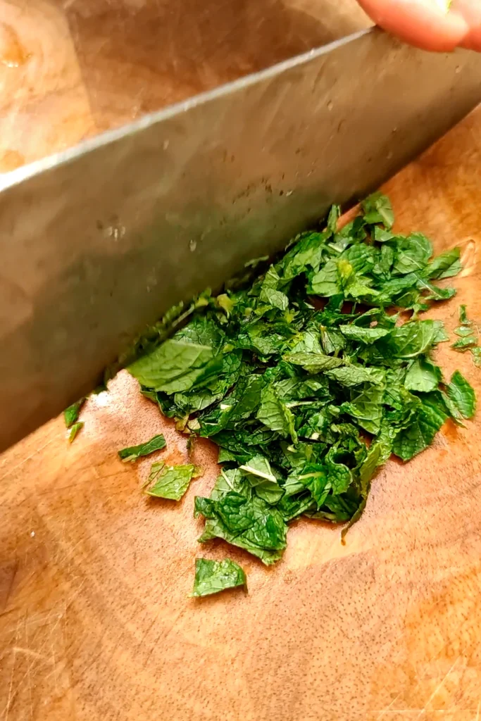 The mint leaves are being chopped with a large knife on a wooden board.