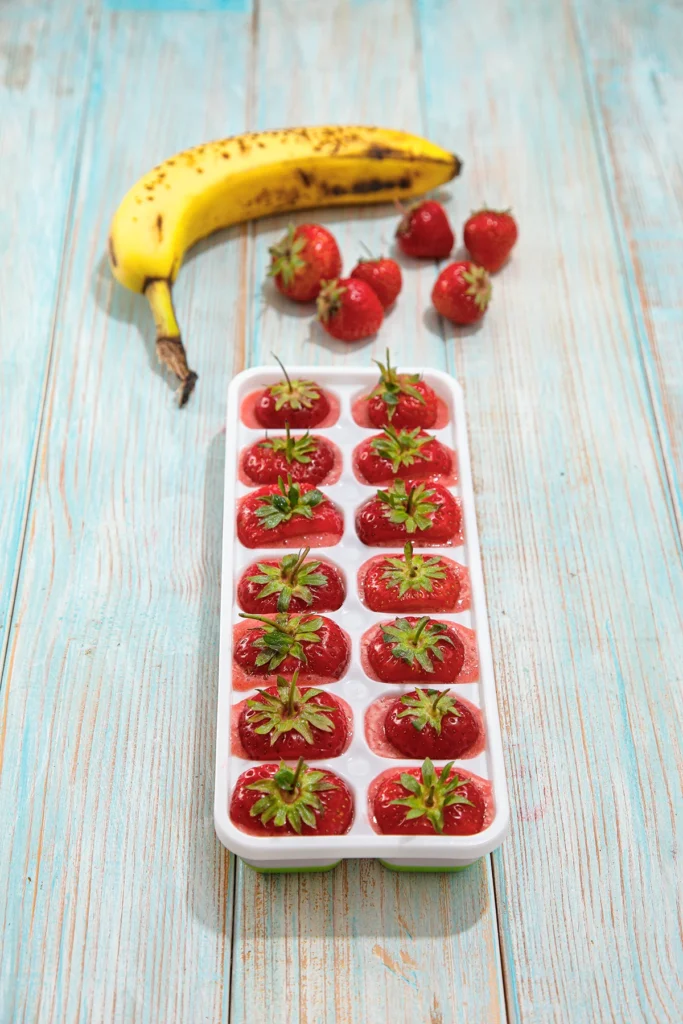 Prepared ice cube mold for strawberry-banana fruit ice cubes stands on a light blue wooden underground. Behind the mold there are a banana and some strawberries.