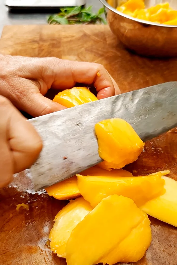 The mango fruit is being chopped with a large knife on a wooden board. In the background stands a bowl with Mango pieces. Behind the board are mint leaves.