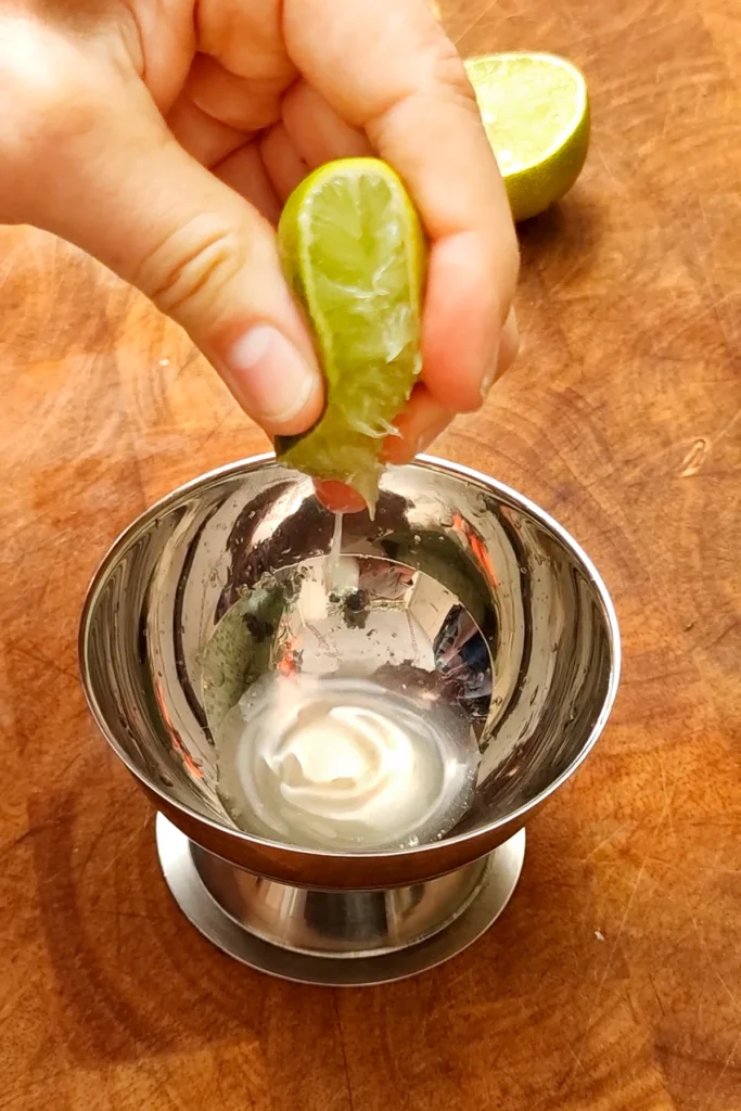 The lime is squeezed with one hand. The juice is collected in a stainless steel bowl which is placed on a wooden board.