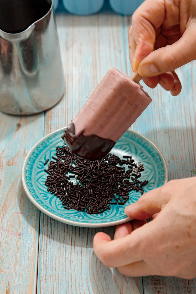 One hand holds a ready-frozen strawberry banana popsicle with a fresh chocolate cap and dips it into the chocolate sprinkles that lies on a plate. In the background is a stainless steel pot with liquid chocolate.