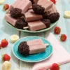 On a turquoise plate is a popsicle with a chocolate cap. Next to it is a pink napkin. Behind it is a large plate with a pile of the same popsicles, surrounded by fresh strawberries and pieces of banana.