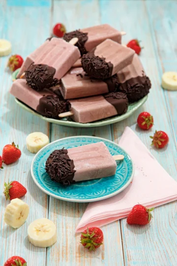 On a turquoise plate is a popsicle with a chocolate cap. Next to it is a pink napkin. Behind it is a large plate with a pile of the same popsicles, surrounded by fresh strawberries and pieces of banana.