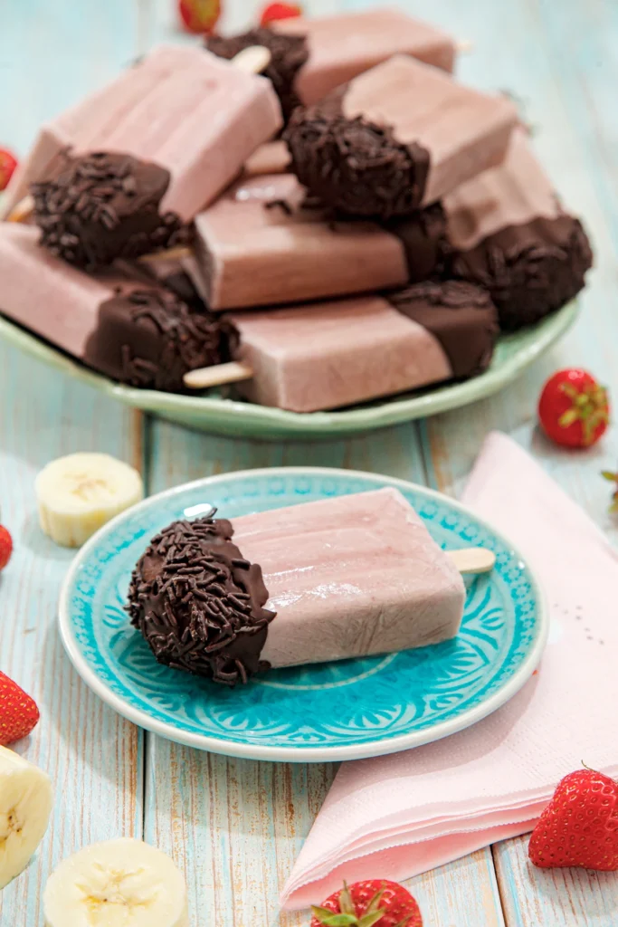 On a turquoise plate is a popsicle with a chocolate cap. Next to it is a pink napkin. Behind it slightly blurred, is a large plate with a pile of the same popsicles, surrounded by fresh strawberries and pieces of banana.