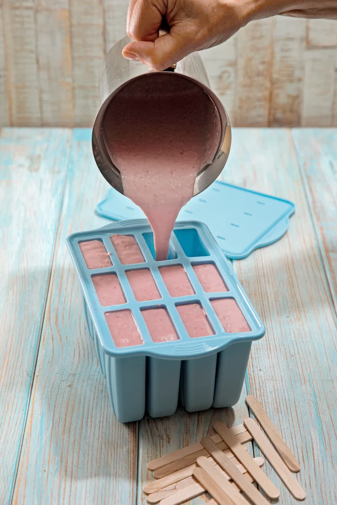 A hand holds a stainless steel pot with strawberry banana puree and pours it into a silicone popsicle mold. In the foreground are wooden popsicle sticks, in the background is the lid for the mold.