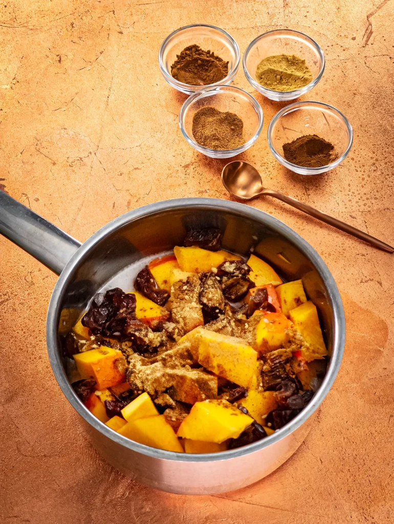 A stainless steel pot with pieces of Hokkaido pumpkin, dates, coconut milk and spices, stands on a bronze-colored base. Behind the pot stand four small glass bowls with spices. A bronze-colored teaspoon lies between the bowls and the pot.