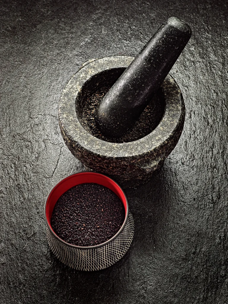 A mortar with pestle and grated black sesame seeds stands on a black slate base. In front of it a bowl with black sesame seeds.