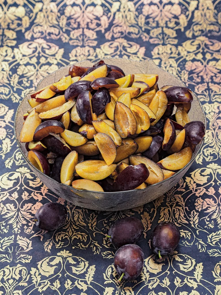 A large glass bowl with quartered plums stands on a patterned surface.