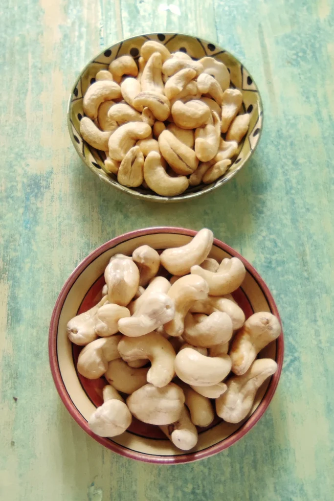 On a light green wooden base are two bowls, in front a pink one with soaked, behind a green one with dried cashew nuts.