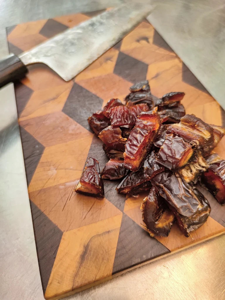 Chopped dates lie on a wooden cutting board. There is a large kitchen knife in the background.