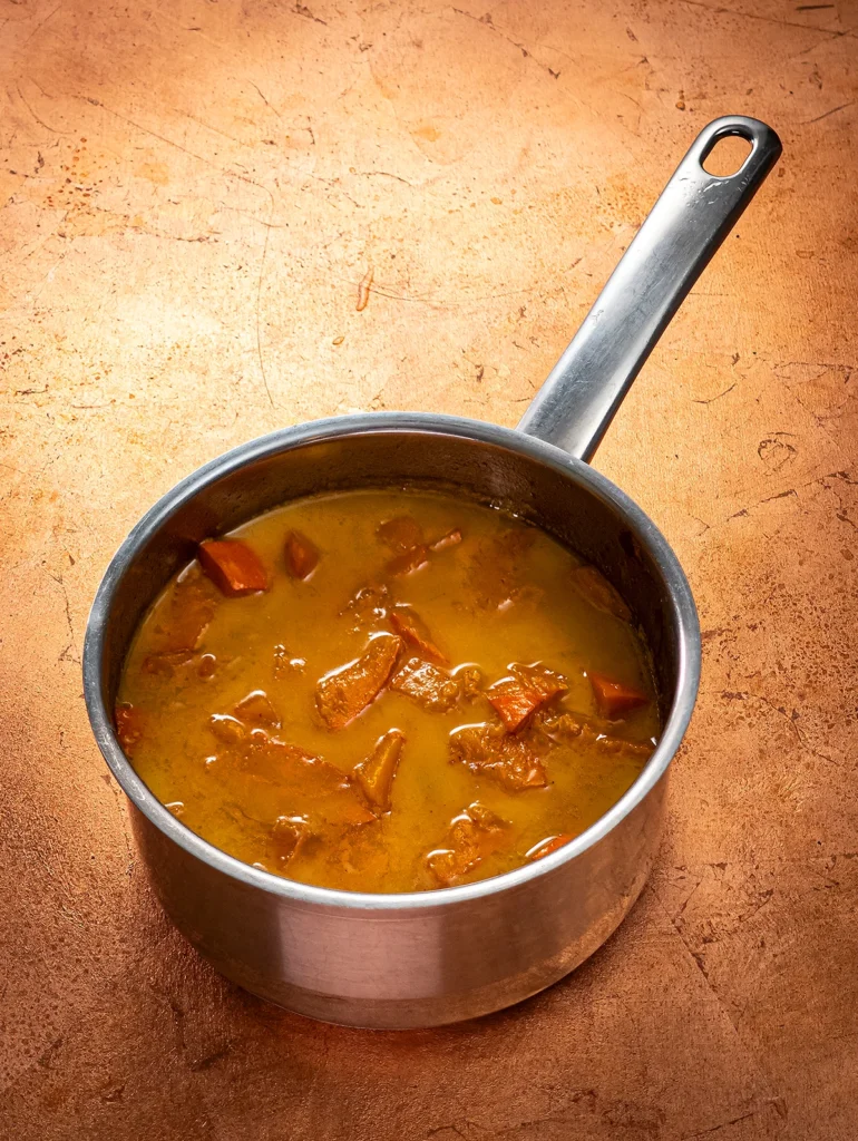 A stainless steel pot with cooked pieces of pumpkin and dates stands on a bronze-colored base.