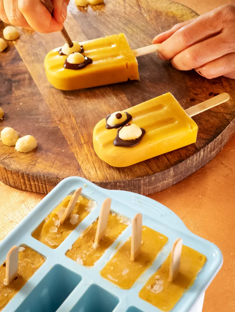 Two ready-frozen and decorated pumpkin popsicles lie on a wooden cutting board. One hand holds a chopstick and adds a chocolate pupil to the macadamia eye. In the foreground you can see the popsicle mold with the other frozen popsicles.