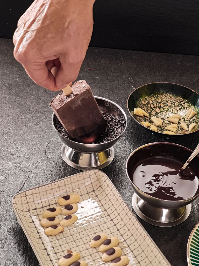 One hand holds a popsicle that has just been dipped in chocolate and is now being dumped into a stainless steel bowl filled with chocolate sprinkles. In the foreground is a flat bowl of halved blanched almonds. Behind it are three bowls of melted chocolate, chocolate sprinkles and almond pieces prepared for the teeth-decoration. Everything stands on a black slate background.