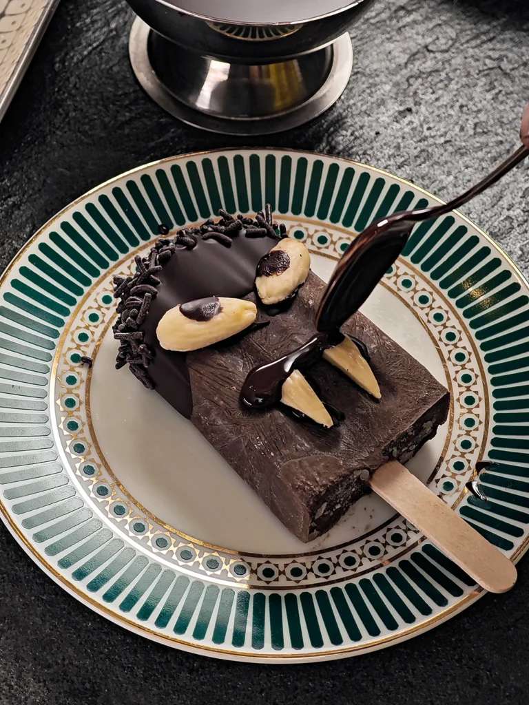 A spoonful of liquid chocolate draws a line on the Halloween popsicle with sesame seeds for the spooky mouth decoration. The Halloween popsicle with sesame seeds is placed on a plate with a border pattern on a slate background.