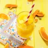 A glass of water with orange turmeric ice cubes and a straw stands on a yellow wooden base. Next to the glass are slices of tumeric, oranges, daisies, and a patterned napkin.