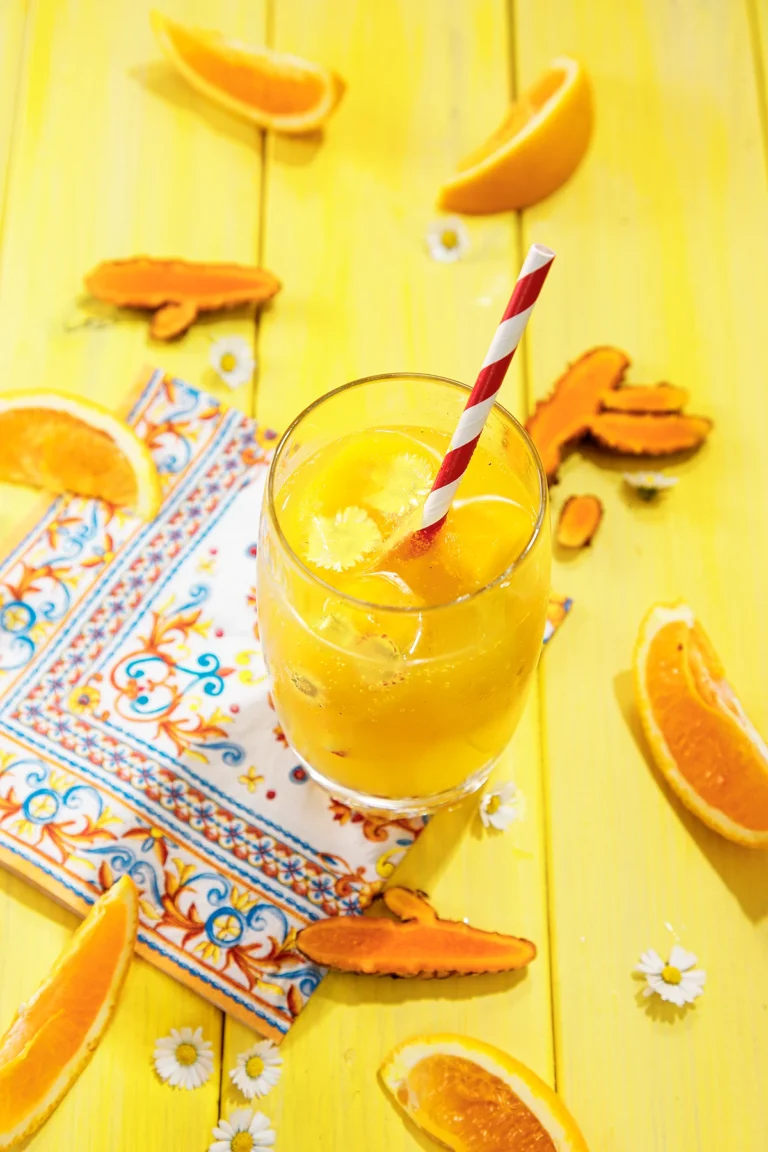 A glass of water with orange turmeric ice cubes and a straw stands on a yellow wooden base. Next to the glass are slices of tumeric, oranges, daisies, and a patterned napkin.