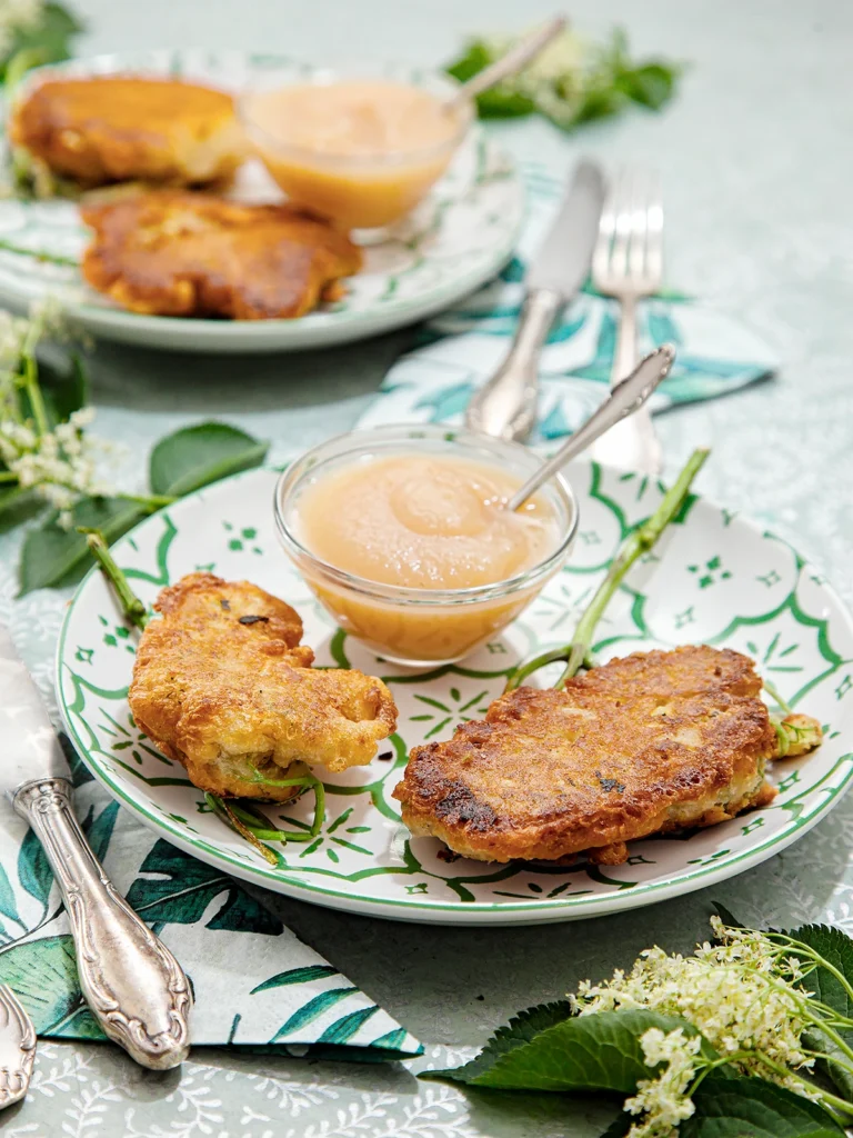 Two elderflower pancakes served on a plate with a little glass bowl of applesauce. There is a napkin and silver cutlery next to the plate. In the background, another plate with the same contents, with a napkin and cutlery, blurred. The plates are placed on a patterned tablecloth with some elder leaves and flowers around them.