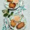 Two elderflower pancakes are served on two plates, each with a little glass bowl of applesauce. There is a napkin and silver cutlery next to the plates. The plates are placed on a patterned tablecloth with some elder leaves and flowers around them.