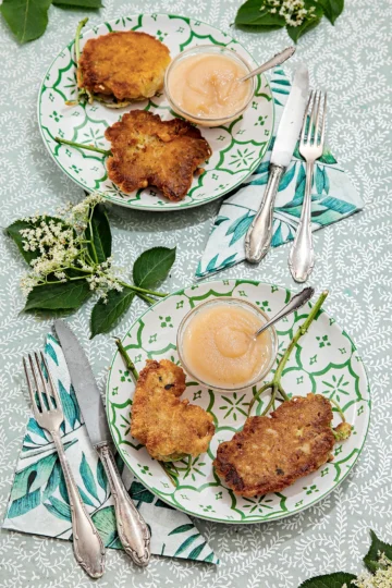 Two elderflower pancakes are served on two plates, each with a little glass bowl of applesauce. There is a napkin and silver cutlery next to the plates. The plates are placed on a patterned tablecloth with some elder leaves and flowers around them.