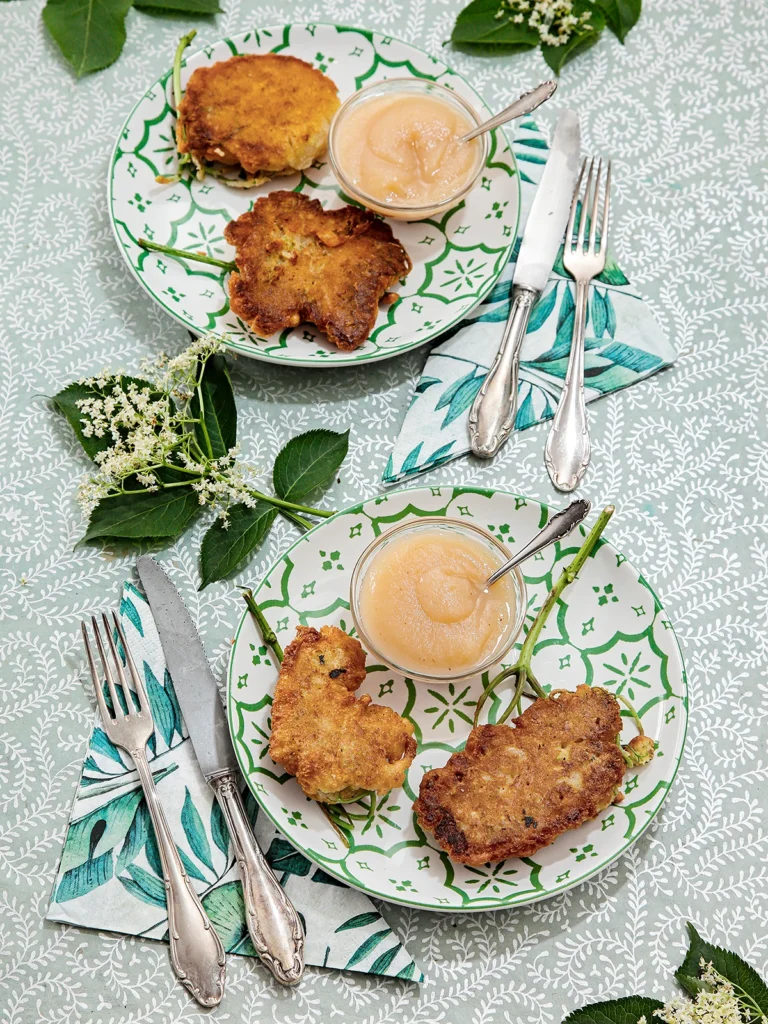 Two elderflower pancakes are served on two plates, each with a little glass bowl of applesauce. There is a napkin and silver cutlery next to the plates. The plates are placed on a patterned tablecloth with some elder leaves and flowers around them.