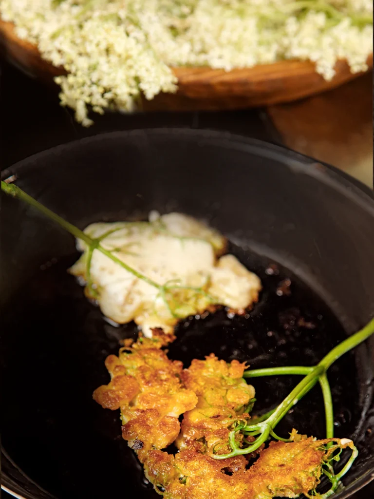 In a black pan there are two elderflower pancakes that are fried in fat. In the background there is a cut round wooden board with elderflowers on it.