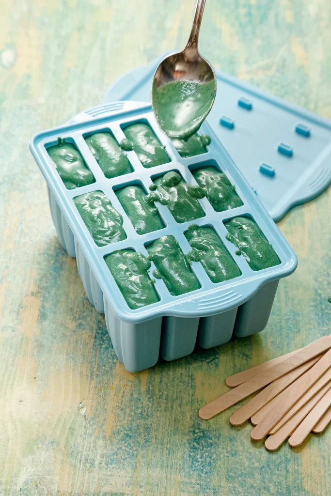 A spoon helps fill the green protein popsicles mixture into a popsicle mold. All compartments are full. Behind the mold is the lid. In front of it are the wooden popsicle sticks. The mold stands on a light green wooden base.