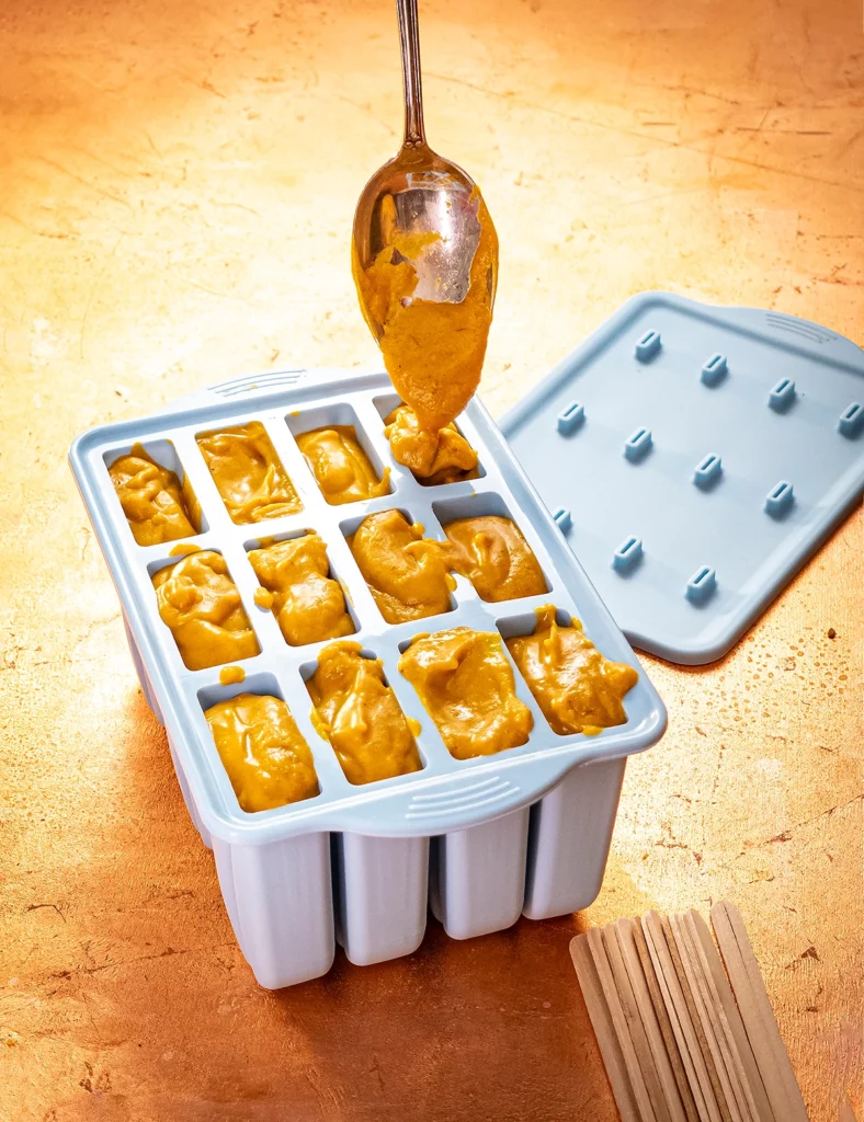 A light blue silicone popsicle mold stands on a bronze-colored background. A spoon helps with the filling, the compartments are all full. In the background is the lid of the mold, in the foreground are the wooden popsicle sticks.