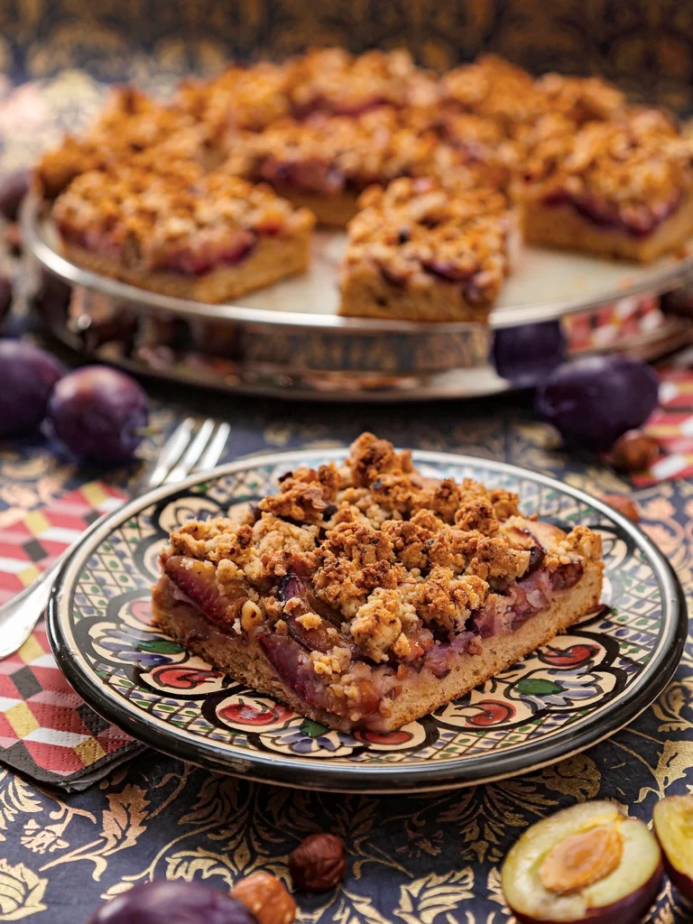 The German plum cake is served on a patterned plate. Attached is a napkin with a cake fork. Behind it, in the blur, a cake serving plate with more pieces of german plum cake. Around it, there are plums and hazelnuts on a patterned surface.