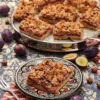 The German plum cake is served on a patterned plate. Attached is a napkin with a cake fork. Behind it a cake serving plate with more pieces of german plum cake. Around it, there are plums and hazelnuts on a patterned surface.