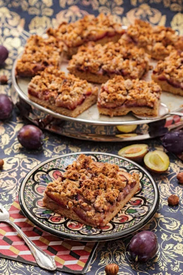 The German plum cake is served on a patterned plate. Attached is a napkin with a cake fork. Behind it a cake serving plate with more pieces of german plum cake. Around it, there are plums and hazelnuts on a patterned surface.