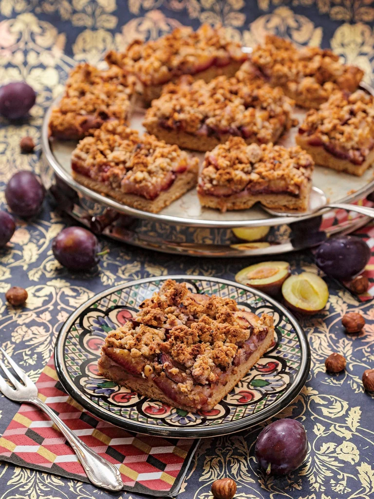 The German plum cake is served on a patterned plate. Attached is a napkin with a cake fork. Behind it a cake serving plate with more pieces of german plum cake. Around it, there are plums and hazelnuts on a patterned surface.