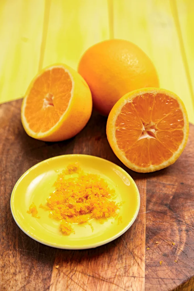 On a round wooden cutting board lies a halved orange, a whole orange and a yellow plate with grated turmeric root. In the background is a yellow wooden background.