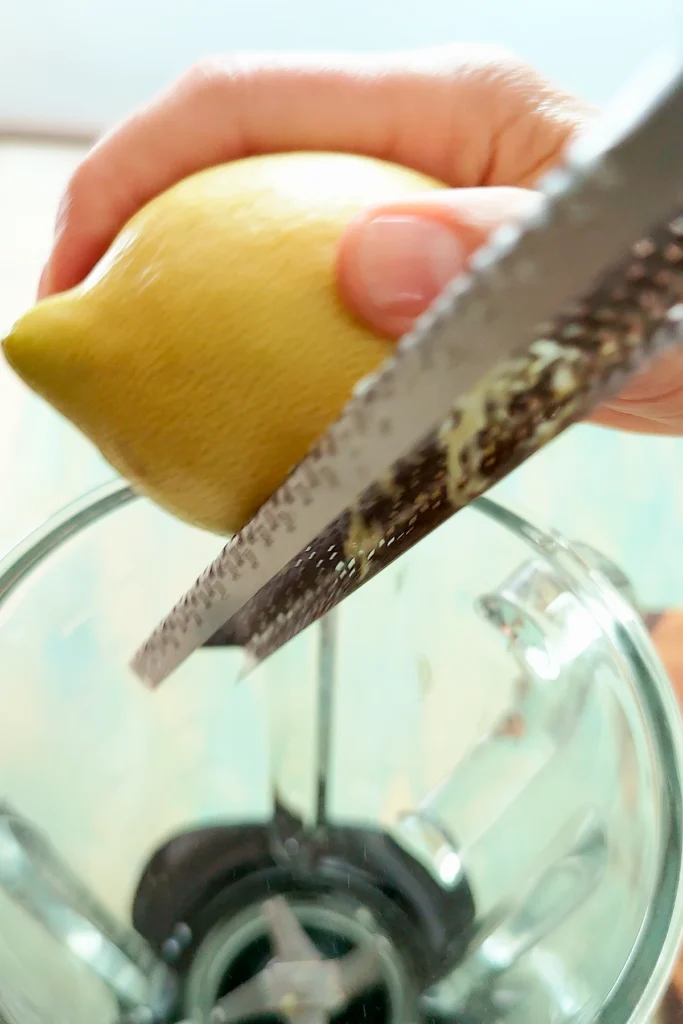 A hand holding a lemon is grating it on a grater above a blender.